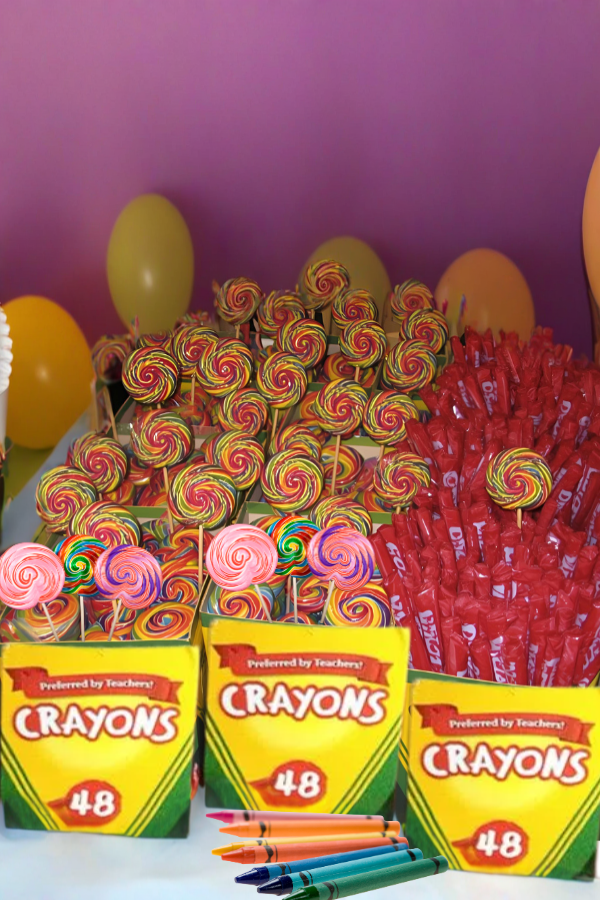 Crayon-themed candy display featuring yellow crayon boxes used as treat holders, filled with rainbow swirl lollipops and red candy sticks, with balloons in the background.