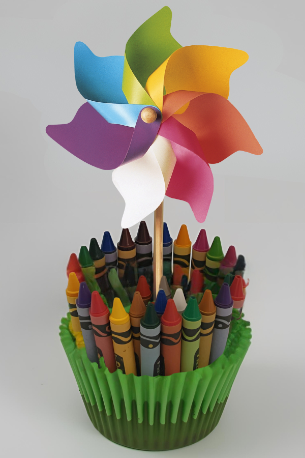 A colorful party centerpiece featuring a rainbow pinwheel placed in a base wrapped with multicolored crayons inside a green cupcake-style holder.