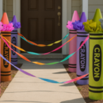 Handmade oversized crayon sculptures in bright modern colors lining an outdoor walkway, connected with ribbons, leading to a front door of a suburban house in daytime.