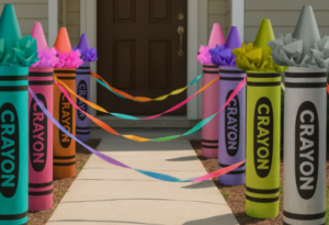 Handmade oversized crayon sculptures in bright modern colors lining an outdoor walkway, connected with ribbons, leading to a front door of a suburban house in daytime.