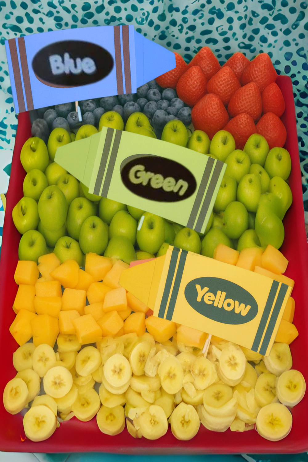 A colorful fruit tray arranged by color with strawberries, blueberries, green apples, cantaloupe, and bananas, each section labeled with a crayon-shaped sign reading Blue, Green, or Yellow.