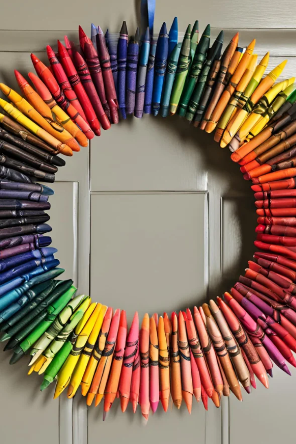 A colorful circular wreath made entirely of crayons arranged in a full rainbow gradient, hanging on a door.
