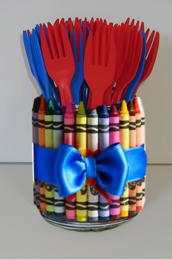 A jar wrapped in multicolored crayons and tied with a bright blue ribbon, filled with red and blue plastic forks.