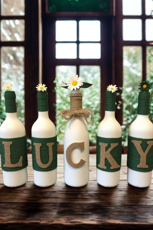 Five handmade decorative bottles wrapped in green twine and burlap letters spelling LUCKY, decorated with white and green flowers