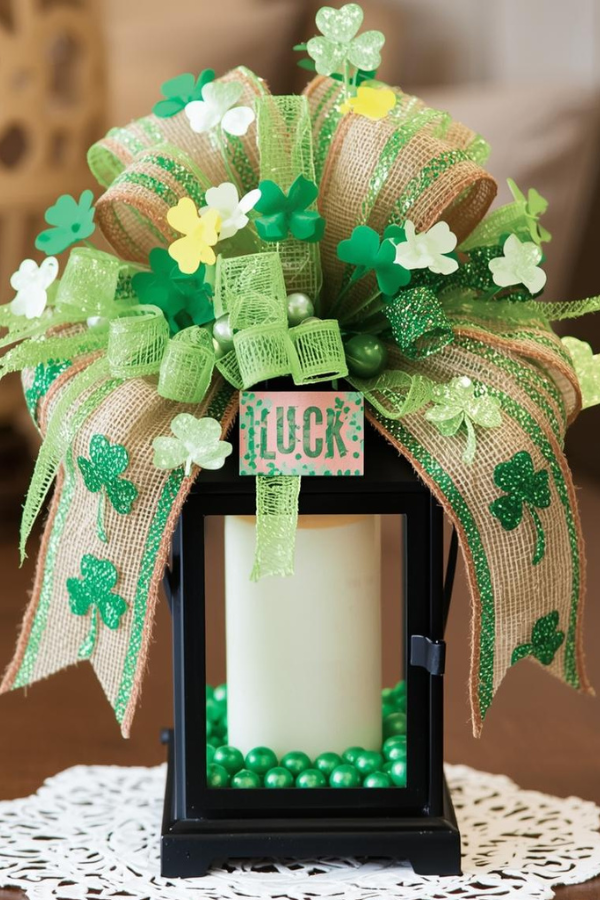St. Patrick’s Day lantern decoration with white candle inside, green beads, burlap bow with shamrocks, glittery green accents, small artificial flowers, and ‘LUCK’ sign on lace doily, festive indoor centerpiece.
