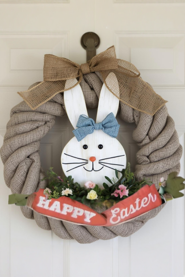 Rustic Easter wreath with a wooden bunny face, burlap bow, blue hair bow, and a “Happy Easter” carrot sign.