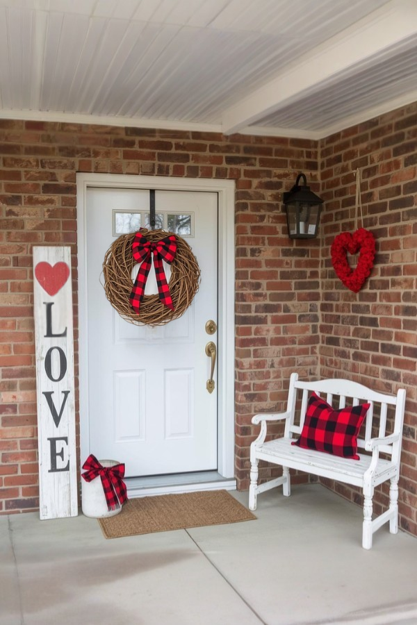 Farmhouse Valentine’s Day front porch with a white door, plaid wreath, LOVE sign, white bench with red and black pillow, and rustic brick walls