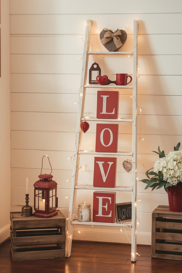 Rustic farmhouse Valentine’s Day ladder decorated with string lights, red LOVE letter blocks, heart accents, lantern, and cozy home décor against a white shiplap wall.