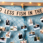 Beach-themed bridal shower photo display featuring a fishing net with clothespins holding couple's photos, a wooden sign reading 'Two Less Fish in the Sea,' and blue fabric draped around for a nautical touch