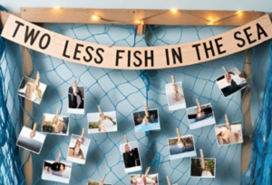 Beach-themed bridal shower photo display featuring a fishing net with clothespins holding couple's photos, a wooden sign reading 'Two Less Fish in the Sea,' and blue fabric draped around for a nautical touch