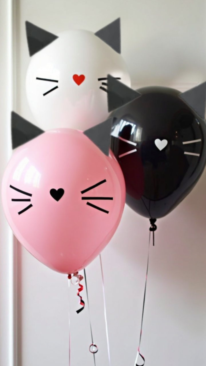 hree cat-themed balloons in pink, white, and black with paper ears, whiskers, and heart-shaped noses, tied with ribbons against a white background