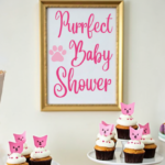 Cat-themed baby shower dessert table with cupcakes decorated with pink cat toppers, a cake, and a framed sign that reads 'Purrfect Baby Shower' in pink script