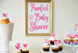 Cat-themed baby shower dessert table with cupcakes decorated with pink cat toppers, a cake, and a framed sign that reads 'Purrfect Baby Shower' in pink script