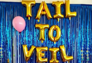 Mermaid bridal shower backdrop with gold balloon letters spelling 'Tail to Veil' against blue shimmering streamers, with a pink balloon decoration