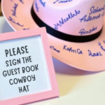 "Pink cowboy hat guest book with handwritten signatures, displayed on a table with a sign that reads 'Please sign the guest book cowboy hat'