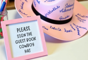 "Pink cowboy hat guest book with handwritten signatures, displayed on a table with a sign that reads 'Please sign the guest book cowboy hat'