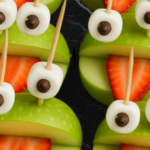Funny green apple frog snacks with strawberry tongues, marshmallow eyes, and chocolate chips on black background