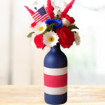 Patriotic centerpiece with red, white, and blue striped bottle vase filled with American flag and festive flowers.