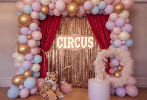 Dream circus theme party backdrop with red velvet curtains, gold tinsel fringe, pastel balloon arch, plush tiger decoration, and a vintage circus sign.