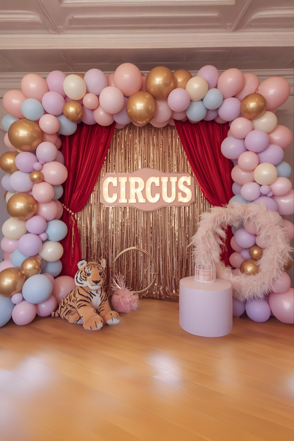 Dream circus theme party backdrop with red velvet curtains, gold tinsel fringe, pastel balloon arch, plush tiger decoration, and a vintage circus sign.