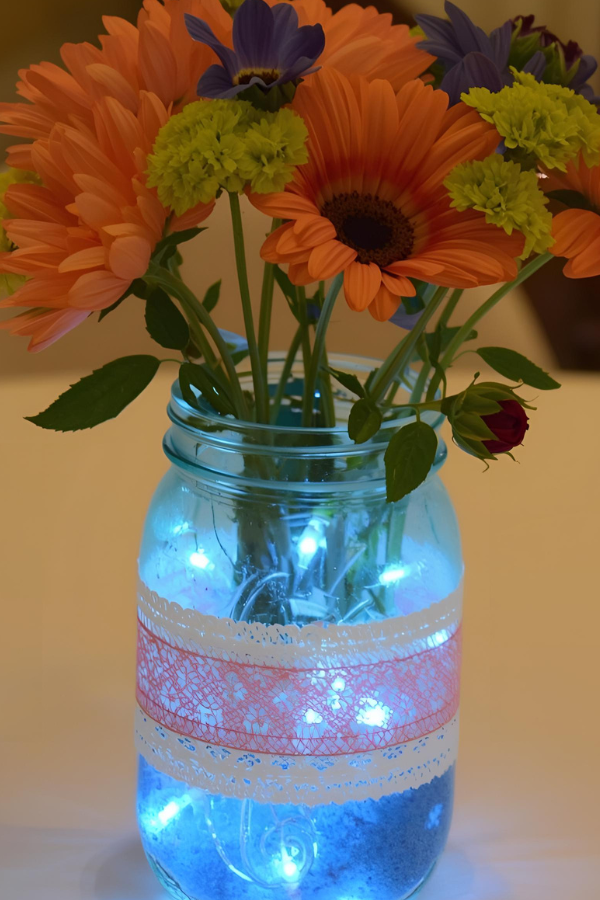 Mason jar centerpiece with orange, yellow, and purple flowers, glowing blue LED fairy lights, and lace ribbon wrapped around the jar.
