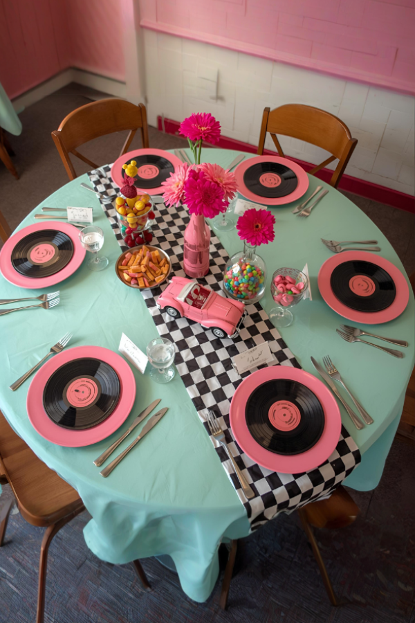 1950s-inspired soda shop party table with a pastel turquoise tablecloth, black-and-white checkered runner, pink plates on vinyl record chargers, a pink toy convertible centerpiece, colorful fruit skewers, candy-filled vases, and light wooden chairs, photographed from a slightly angled overhead perspective.