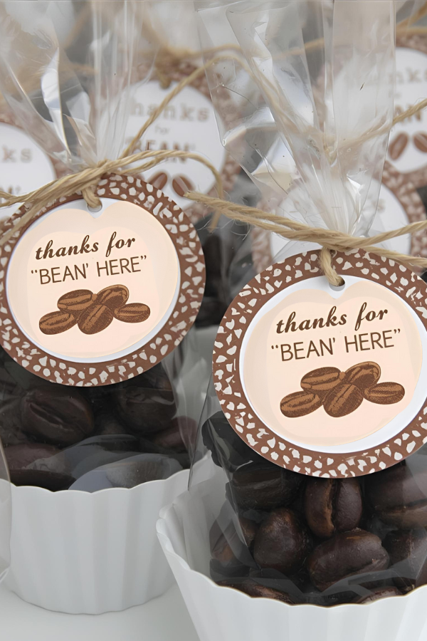 Coffee-themed party favor bags filled with roasted coffee beans and tied with twine, decorated with round tags that say 'Thanks for BEAN' HERE' in a warm café-inspired design.