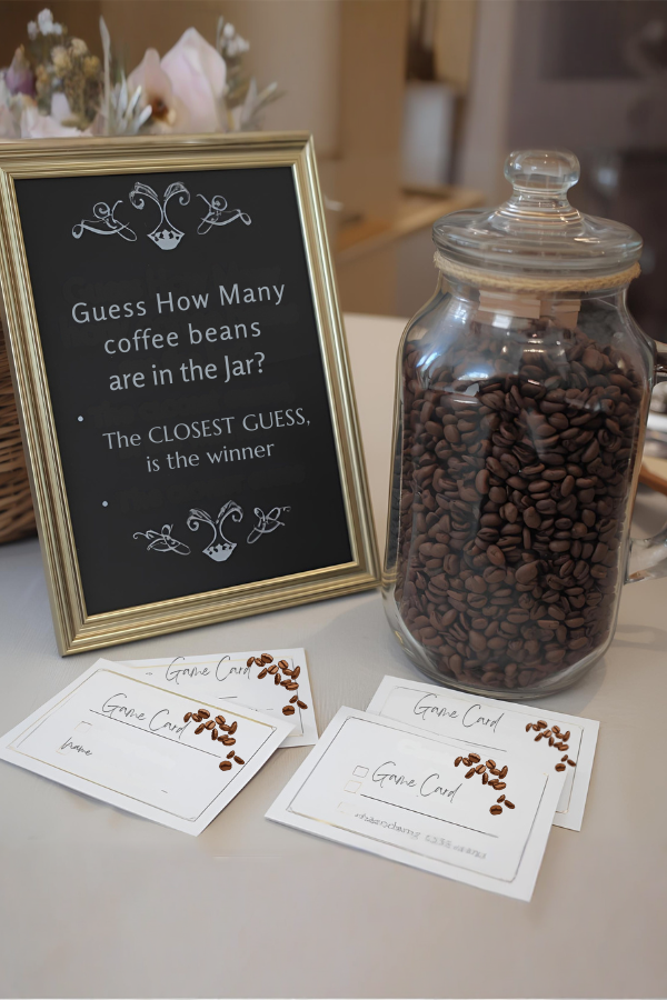 A glass jar filled with coffee beans beside a framed sign inviting guests to guess how many beans are inside, with game cards laid out on the table.