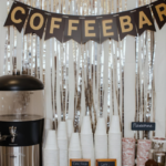 A self-serve coffee bar decorated with a COFFEEBAR banner, stacked cups, a large coffee dispenser, flavor syrups, and stirrers arranged on a white dresser with a cozy café-style backdrop.