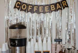 A self-serve coffee bar decorated with a COFFEEBAR banner, stacked cups, a large coffee dispenser, flavor syrups, and stirrers arranged on a white dresser with a cozy café-style backdrop.