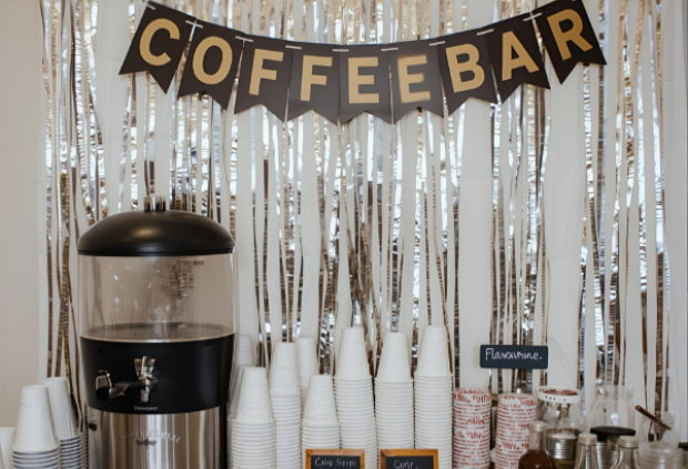 A self-serve coffee bar decorated with a COFFEEBAR banner, stacked cups, a large coffee dispenser, flavor syrups, and stirrers arranged on a white dresser with a cozy café-style backdrop.