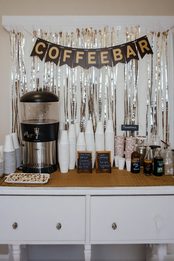 A self-serve coffee bar decorated with a COFFEEBAR banner, stacked cups, a large coffee dispenser, flavor syrups, and stirrers arranged on a white dresser with a cozy café-style backdrop.