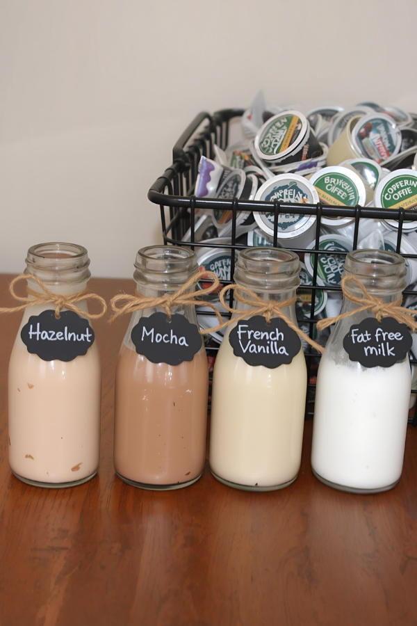 "Glass bottles labeled with hazelnut, mocha, French vanilla, and fat-free milk arranged in front of a basket of assorted coffee pods, creating a cozy café-style drink station.