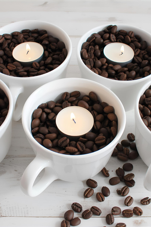 White ceramic mugs filled with roasted coffee beans and topped with small lit tealight candles, styled as cozy café-themed centerpieces.