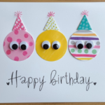 Handmade birthday card with colorful paper circles featuring googly eyes and hand-drawn party hats on white cardstock.