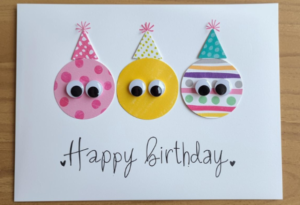 Handmade birthday card with colorful paper circles featuring googly eyes and hand-drawn party hats on white cardstock.