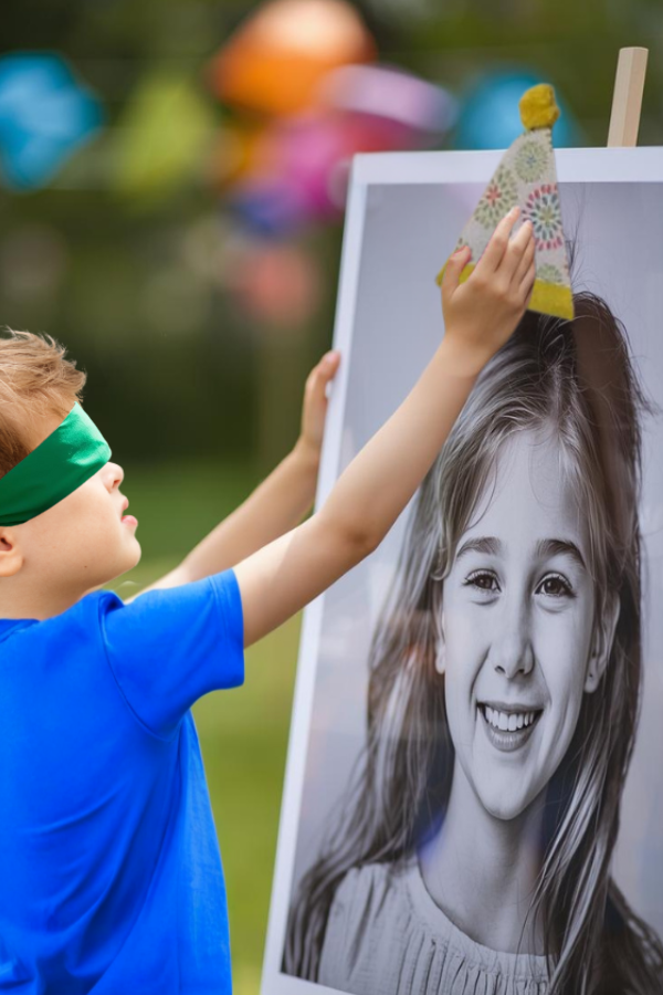 Child playing a blindfolded party game by placing a hat on a photo at an outdoor birthday party.