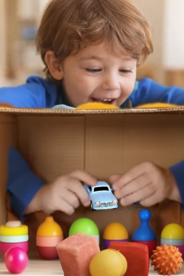 Preschool-aged child playing a “What’s in the Box?” sensory guessing game, reaching through a decorated cardboard box to explore textured objects indoors.