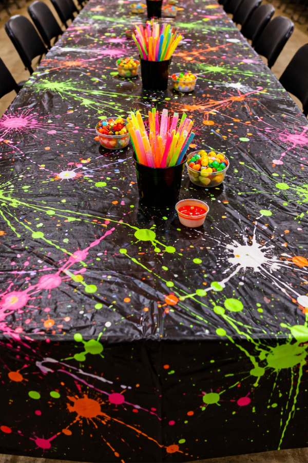 Long party table with black tablecloth covered in bright neon paint splatters and chairs on both sides.