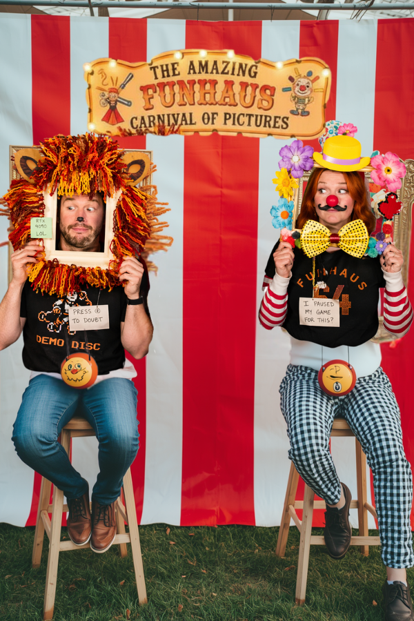 Colorful DIY carnival photo booth with red-and-white striped backdrop, two people holding whimsical costume frames (lion and clown) with goofy props, playful lighting, and chaotic handmade decorations.