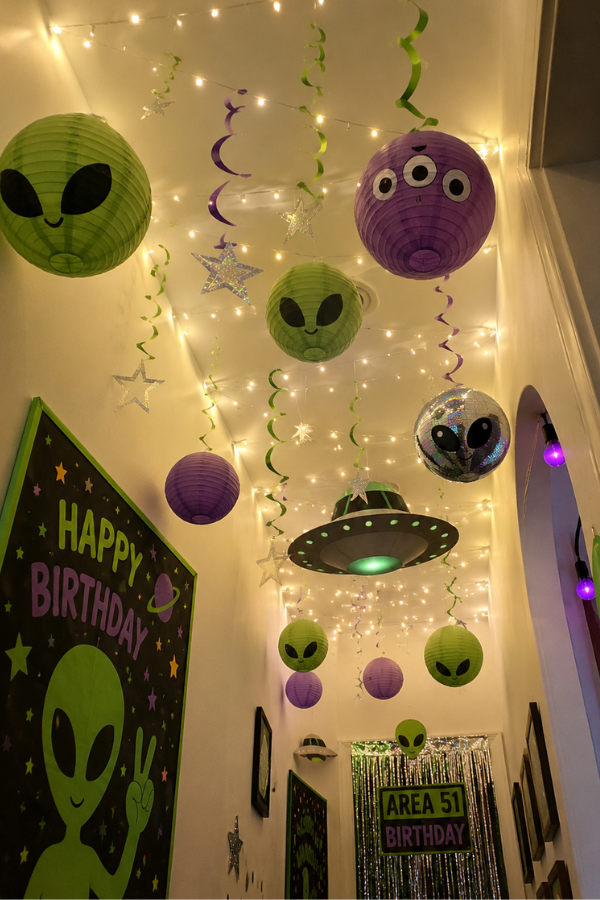 Alien-themed birthday party hallway with glowing fairy lights, hanging paper lantern aliens, UFO decorations, and neon green and purple space-themed decor.